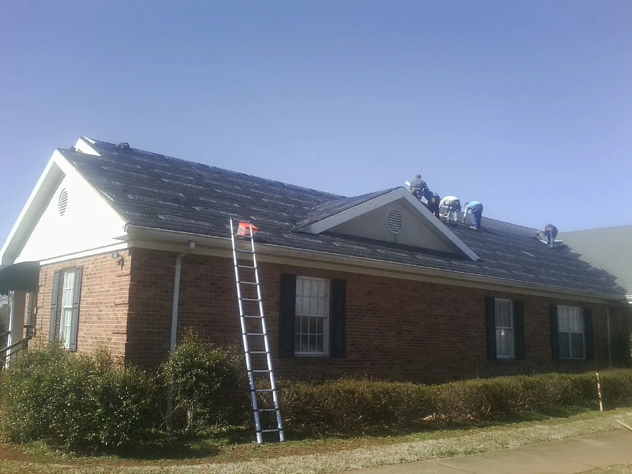 Crew tearing shingles off a brick ranch roof with a ladder set — Metal Roof Repair work by our crew in Grafton